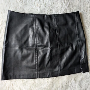 Svart skinnkjol från Monki - Säljer en svart kort skinnkjol från Monki i storlek 42. Kjolen har en clean look med markerade sömmar framtill och dragkedja i sidan. Perfekt för dig som gillar en edgy och stilren vibe.