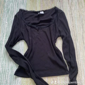 Svart ribbad topp med spets från H&M - Svart långärmad topp från H&M Divided i ribbad bomull med söt spetskant och liten rosett vid halsringningen. Sitter snyggt och har en feminin vibe, perfekt till jeans eller kjol. Storlek XS.