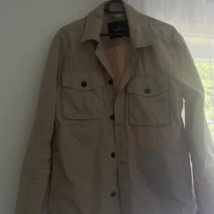 Beige overshirt från Morris - Snygg beige overshirt från Morris med klassisk krage och två stora bröstfickor med knappar. Långärmad modell i bomull, perfekt att slänga över en t-shirt. Stora knappar framtill och diskret broderad detalj på ena fickan ger en clean look.