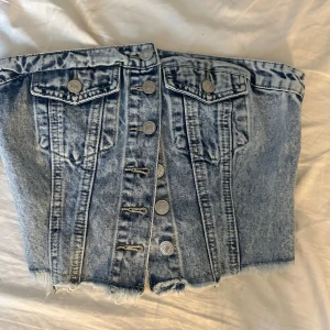 Jeans tubtopp från SHEIN med knappar - Säljer en snygg tubtopp i ljusblå denim från SHEIN. Toppen har coola silverfärgade knappar framtill. Baksidan har smock för extra stretch och passform. Perfekt för dig som gillar streetstyle och denim vibes.