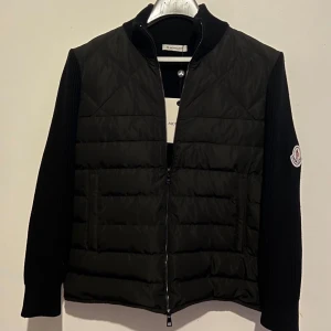 Svart pufferjacka från Moncler - Svart pufferjacka från Moncler med quiltad front i syntetmaterial och stickade ärmar och rygg. Jackan har dragkedja framtill, två fickor och Moncler-logga på ärmen. Perfekt för dig som vill ha en stilren och sportig look.