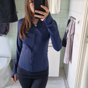 Mörkblå sportig vindjacka - Säljer en mörkblå, figursydd vindjacka med dragkedja framtill och hög krage. Jackan är långärmad och tillverkad i ett slätt, syntetiskt material som passar perfekt för träning eller aktiva dagar. Enkel och stilren design utan mönster eller extra detaljer.