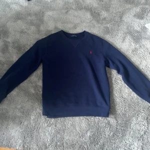 Mörkblå sweatshirt från Polo Ralph Lauren - Klassisk mörkblå sweatshirt från Polo Ralph Lauren med rund hals och röd broderad logga på bröstet. Tröjan har långa ärmar och är tillverkad i mjuk bomull, perfekt för en chill och stilren look.