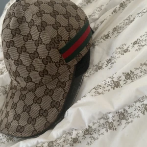 Gucci keps med GG-mönster och röd/grön detalj - Säljer en ikonisk Gucci keps i beige och brunt GG-monogram med klassisk röd och grön rand på sidan. Kepsen har justerbart spänne bak och mesh-detaljer på sidorna. Tillverkad i canvas med läderdetaljer och snygg formad skärm. Perfekt för dig som vill sticka ut. Skick 7,8 av 10 nypris ligger runt 5500kr uppåt har sett en hemsida som säljer den för 9 tusen.