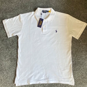 Vit pikétröja från Polo Ralph Lauren - Klassisk vit pikétröja från Polo Ralph Lauren med korta ärmar och broderad logga i lila på bröstet. Tröjan har krage och knäppning med två knappar framtill. Tillverkad i mjuk bomull för en clean och fräsch look.