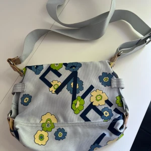 Blommig axelväska från Benetton - Snygg axelväska i ljusblått från United Colors of Benetton med blommigt mönster i blått, grönt och gult. Väskan har justerbar rem, detaljer i skinn och dragkedja upptill. Perfekt storlek för det viktigaste och en cool detalj till din outfit.