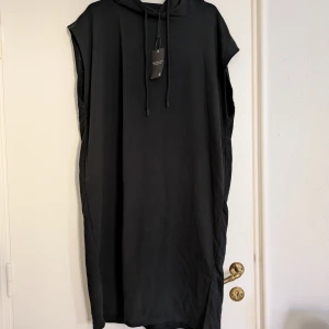 Svart hoodieklänning - Svart ärmlös hoodieklänning från i sweatshirttyg. Klänningen har huva med snörning, rak passform och slits på vardera sida. Perfekt för en chill och avslappnad stil. Oversize.