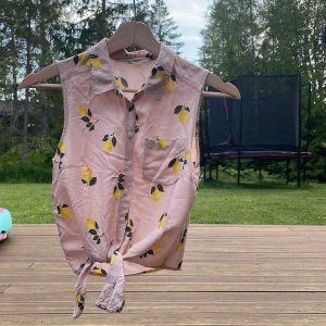 Rosa ärmlös blus med citronmönster - Söt rosa ärmlös blus med krage och knappar framtill. Toppen har ett fräscht citronmönster i gult och grönt, en liten bröstficka och knytning nedtill för en chill sommarlook. Perfekt för dig som vill sticka ut med färg och mönster!                                                             Den är lite liten i storleken 