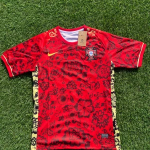 Portugal Nike fotbollströja röd blommig - Portugal fotbollströja från Nike i rött med svarta blommiga mönster och gula detaljer. Kortärmad modell i lätt och ventilerande Dri-FIT-material. Broderad Nike-logga och Portugals landslagsemblem på bröstet. Unik design med blommor och kontrasterande sidor.