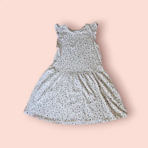 Prickig ljus kortklänning med volang - Supersöt ljus kortklänning med små prickar i svart, rosa och blått. Klänningen har volangdetaljer vid axlarna och är ärmlös. Den har en rund halsringning och ett mjukt, flowy fall. Perfekt för varma dagar och enkel att styla med sneakers eller sandaler.