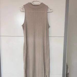 Beige ribbad ärmlös långklänning -  Beige, ribbad långklänning med hög hals och slits på sidorna. Klänningen är ärmlös och har en clean, minimalistisk look som är superenkel att styla.
