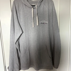 Grå hoodie från Lexington - Snygg grå hoodie från Lexington med broderad logga på bröstet. Tröjan har huva med snörning, känguruficka och långa ärmar. Perfekt för chill dagar och enkel att matcha med jeans eller mjukisbyxor.