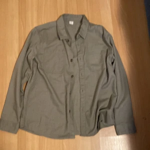 Grå overshirt med bröstfickor - Snygg grå overshirt med klassisk krage och två bröstfickor. Skjortan har knappar framtill där det står 'Since 1985' och är tillverkad i ett mjukt bomullstyg. Perfekt att bära öppen över en t-shirt eller stängd för en mer clean look.