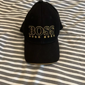 Svart keps från Hugo Boss med gulddetaljer - Snygg svart keps från Hugo Boss med broderad BOSS-logga i gult framtill. Justerbar rem baktill med metallspänne och gul rand. Klassisk böjd skärm med gul kant. Perfekt accessoar för en stilren look.