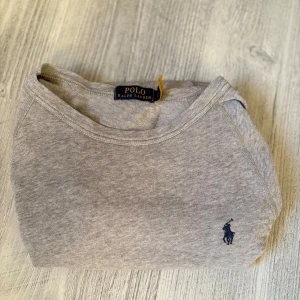 Grå långärmad tröja Polo Ralph Lauren - Snygg grå långärmad tröja från Polo Ralph Lauren med klassisk rund hals och den ikoniska blå loggan broderad på bröstet. Tillverkad i mjuk bomull som känns skön mot huden. Perfekt basic-plagg att ha i garderoben. Qr kod är på tröjan som visar äktenskapet!