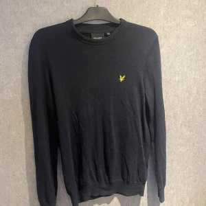 Mörkblå marineull tröja från Lyle & Scott - Svart stickad tröja från Lyle & Scott med klassisk rund hals och gul broderad logga på bröstet. Tröjan har lång ärm och en enkel, clean look som funkar till allt. Perfekt för dig som gillar stilrena plagg med en touch av street.