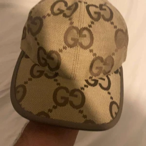 Beige keps från Gucci med GG-mönster - Snygg beige keps från Gucci med det klassiska GG-monogrammet i brunt över hela kepsen. Justerbar rem baktill med metallspänne. Tillverkad i tåligt canvasmaterial och har en böjd skärm. Perfekt för dig som vill sticka ut med en lyxig accessoar.