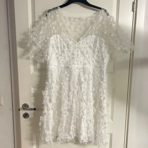 Studentklänning  - Superfin vit klänning med 3D-blommor och transparent spets över axlar och ärmar. Klänningen har v-ringning och söt, luftig känsla. Perfekt för sommarens alla tillfällen när du vill sticka ut lite extra, exempelvis studenten. Helt ny enbart testad en gång, dock utan prislapp. Nypris: 1100kr