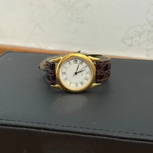 Apollo guldig klocka med läderband - Stilren klocka från Apollo med rund, guldig boett och vit urtavla med romerska siffror. Armbandet är brunt och gjort i äkta läder med krokopräglat mönster. Klassisk design som passar dig som gillar tidlösa accessoarer.