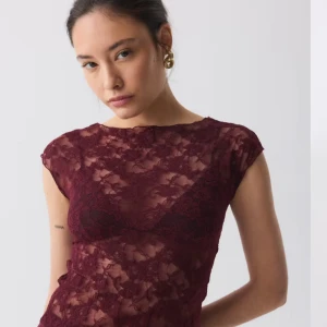 Vinröd spetsblus från Gina Tricot - Säljer en vinröd blus i genombruten spets från Gina Tricot. Modellen är figurnära med korta ärmar och rund halsringning. Blusen har ett romantiskt blommönster i spetsen och är perfekt för att skapa en trendig och feminin look.