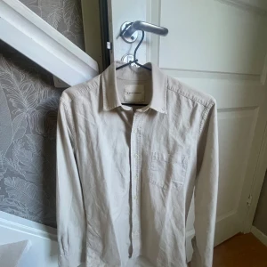 Beige skjorta från A Day's March - Stilren beige skjorta från A Day's March i ekologisk bomull. Klassisk krage, bröstficka och knäppning framtill. Skjortan har lång ärm och en clean look som passar till många olika outfits. Ej strykt på bilderna, använd 2-3 gånger så i gott skick. Nypris:1150kr