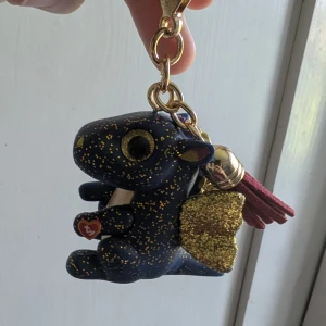 Nyckelring med blå drake och glitter - Supergullig nyckelring från Lekia i form av en blå drake med guldglittriga vingar och detaljer. Draken har stora ögon, guldprickar och en vinröd tofs. Perfekt att hänga på väskan eller använda till nycklarna.