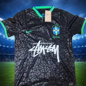 Svart Brasil x Stüssy x Nike fotbollströja - Unik svart Brasilien fotbollströja i samarbete med Stüssy och Nike. Tröjan har korta ärmar, gröna detaljer vid krage och ärmslut, samt ett subtilt leopardmönster över hela tyget. Stor Stüssy-logga framtill och Brasilien-emblem på bröstet. Perfekt för dig som gillar streetstyle och fotboll Storlek S.M.