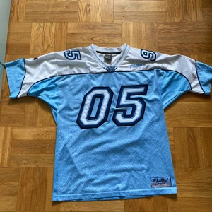 FUBU blå och vit fotbollströja 05 - Retro FUBU fotbollströja i meshmaterial med stora siffror 05 fram och bak. Tröjan är ljusblå med vita partier upptill och på ärmarna, samt mörkblå detaljer och broderade patches. Oversized passform och korta ärmar, perfekt för sportig streetstyle.                                      Kan mötas upp i Umeå 