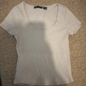 Ljusblå ribbad topp från 157 - ( jag säger M) En ljusblå ribbad topp från 157 med korta ärmar och rundad, djup urringning. T-shirten har en slim passform och är tillverkad i ett mjukt, stretchigt material som sitter snyggt på kroppen. Perfekt att styla med jeans eller kjol.