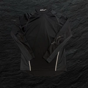 Svart långärmad tröja Bauer - Svart långärmad träningströja från Bauer med halsskydd och diskreta grå mönsterdetaljer på ärmar och axlar. Tröjan är tillverkad i ett syntetmaterial som andas, perfekt för intensiva hockeymatcher/träningar.