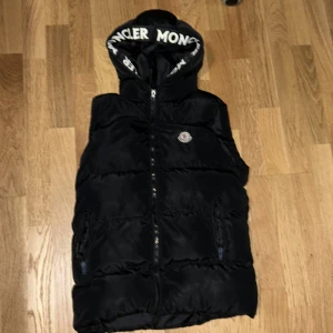 Svart dunväst från Moncler med huva - Svart dunväst från Moncler med stor vadderad huva och vit Moncler-logga broderad på bröstet. Västen har dragkedja framtill, två fickor med dragkedja och coolt Moncler-tryck på huvkanten. Perfekt för lager-på-lager och streetstyle.
