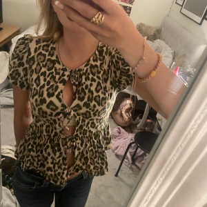 Leopardmönstrad blus från Gina Tricot - Supersnygg blus med leopardmönster från Gina Tricot, använd cirka en gång och är i jätte bra skick. Modellen har puffiga korta ärmar, volang nertill och tre knytningar framtill som ger en cool och trendig look. Perfekt för dig som vill sticka ut med ett djurmönstrat statement-plagg.