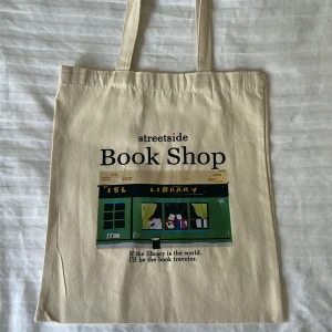Beige tygkasse med Book Shop-tryck - Tygkasse i beige bomull med tryck av en grön bokhandel och texten 'streetside Book Shop'. Perfekt för böcker, laptop eller shopping. Långa handtag gör den enkel att bära på axeln. 