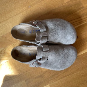 Birkenstock Boston Mocka Taupe - Birkenstock Boston i taupe mocka med rund tå och justerbart spänne i metall. Skorna har en platt sula och är perfekta för en chill och avslappnad stil. Klassisk slip-in modell med mjuk fotbädd för extra komfort.