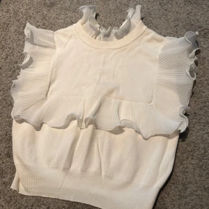 Vit volangblus från Zara - Supersöt vit blus från Zara med plisserade volanger längs axlar och hals. Hög krage och ärmlös design ger en trendig look. Perfekt att styla med jeans eller kjol för en chic vibe.
