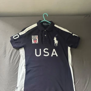 Mörkblå Ralph Lauren USA pikétröja - Snygg mörkblå “chief keef” pikétröja från Ralph Lauren med vita detaljer och stora USA-tryck fram och bak. Klassisk polokrage, korta ärmar och broderad polospelare på bröstet. Perfekt för dig som gillar sportig och preppy stil.