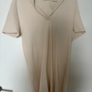 Beige kort klänning från Zara - Enkel och stilren beige kortklänning från Zara med v-ringning och korta ärmar. Klänningen har en mjuk känsla och är tillverkad i bomull, perfekt för en avslappnad look. Snygg krage som ger en modern touch.