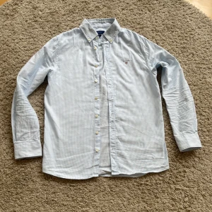Ljusblå randig skjorta från GANT - Snygg ljusblå skjorta från GANT med tunna vita ränder och klassisk button down-krage. Skjortan har broderad GANT-logga på bröstet, långa ärmar och knäppning framtill. Tillverkad i mjuk bomull för en skön känsla.