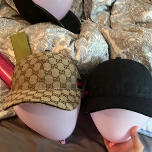 Båda keps snygg  - Två snygga  kepsar – en i beige med klassiskt -mönster och en i svart med diskret -mönster. Båda har den ikoniska gröna och röda randen på sidan och justerbar rem bak. Perfekt accessoar för att lyfta din stil.