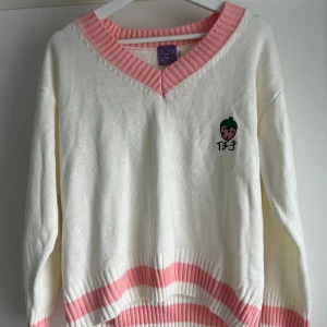 Vit och rosa stickad tröja Honey Bomber - Supersöt vit stickad tröja från Honey Bomber med breda rosa ribbade kanter och v-ringning. Broderad jordgubbe med japansk text på bröstet. Långärmad och med mjuk, lite loose passform. Perfekt för dig som gillar kawaii-stil och pastellfärger.