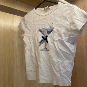 Vit topp med martiniglas från Subdued - Vit kortärmad babytee topp från Subdued med tryck av ett martiniglas och en liten blå rosett framtill. T-shirten har en klassisk rund halsringning och är gjord i mjukt bomullsmaterial. Perfekt för dig som gillar unika detaljer och coola prints. Stl M men liten i storlek och skulle passa S/XS bättre