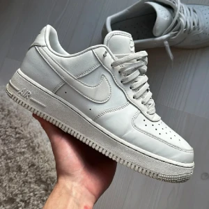 Air Force 1 - Klassiska Nike Air Force 1 sneakers i helvitt skinn med perforerad tå och robust sula. Snygg low-top modell med snörning och ikonisk Swoosh på sidorna. Skorna är tillverkade i mjukt, andningsbart läder och passar perfekt till streetwear. 