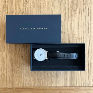 Daniel Wellington klocka med svart läderband - Stilren klocka från Daniel Wellington med vit urtavla, romerska siffror och svart läderband. Batteri behöver bytas. Nypris: 1999 kr