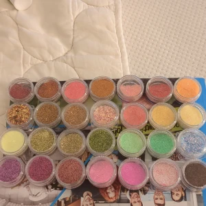 Set med 28 burkar nagelglitter - Säljer ett set med 28 små burkar nagelglitter i olika färger som rosa, guld, grönt, lila, orange, gult och blått. Helt oanvända