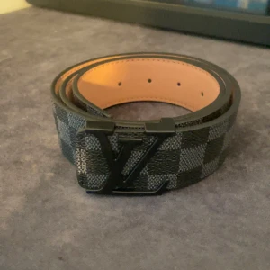 Louis Vuitton rutigt svart bälte - Snyggt bälte från Louis Vuitton med klassiskt rutigt mönster i svart och grått. Bältet har en stor LV-logga i svart metall som spänne och är gjort i skinn med en lyxig känsla. Perfekt accessoar för att lyfta din outfit.