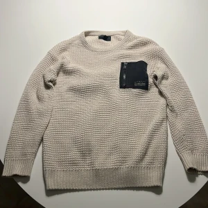 Beige stickad tröja D.M.D med ficka - Beige stickad tröja från D.M.D by denim day med lång ärm och cool svart bröstficka med dragkedja. Tröjan har grovstickad struktur och ribbade muddar vid ärmslut och nederkant. Perfekt för dig som gillar en enkel men ändå unik stil.