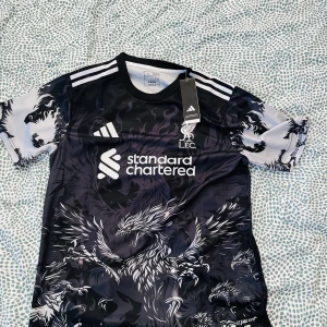 Liverpool x Adidas svart draktröja - Snygg Liverpool fotbollströja från Adidas med svart och vit drakdesign över hela tröjan. Kortärmad modell med vita axelränder och klubbmärke samt sponsortryck på bröstet. Tillverkad i lätt och ventilerande material, perfekt för match eller träning.