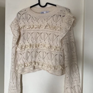 volangtröja Zara - Supersöt beige stickad tröja från Zara med volangdetaljer och hålmönster. Tröjan har lång ärm, rund halsringning och dekorativa virkade ränder framtill. 