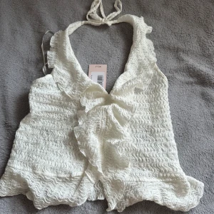 Vit volang halterneck topp från Nelly - Supersöt vit halterneck topp från Nelly med volangdetaljer och vågig struktur. Toppen knyts i nacken och har en omlottkänsla framtill. Perfekt för dig som gillar romantiska och trendiga plagg med extra detaljer.