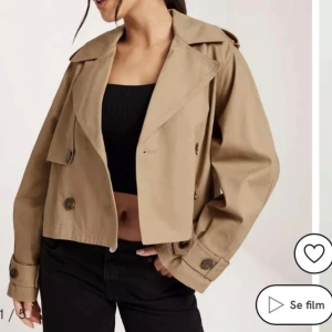 Beige croppad trenchcoat - Snygg croppad trenchcoat i beige med bred krage och stora knappar. Jackan har långa ärmar med knäppning vid ärmslut och klassiska trench-detaljer. Perfekt för dig som vill ha en trendig och stilren look till våren eller hösten. Nypris 600kr aldrig använd 